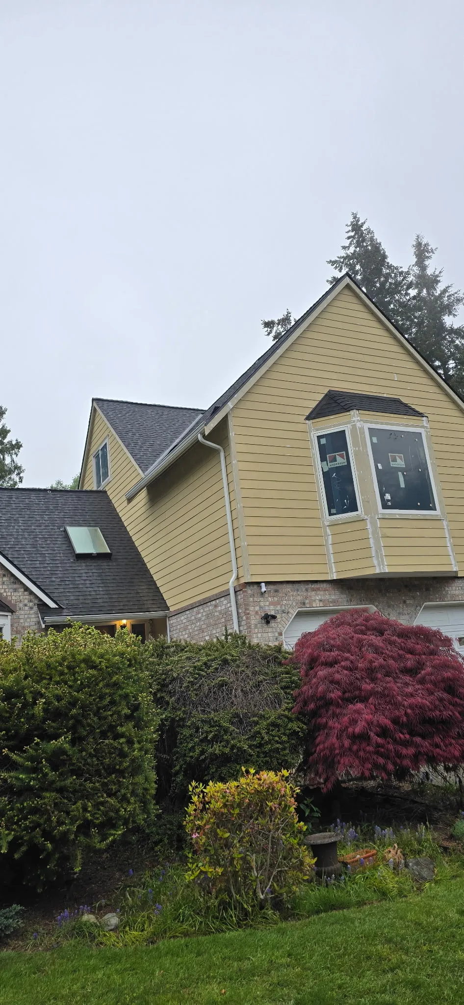 Yellow lap siding on a two-story home with a bay window and brick base, trees and shrubs in front