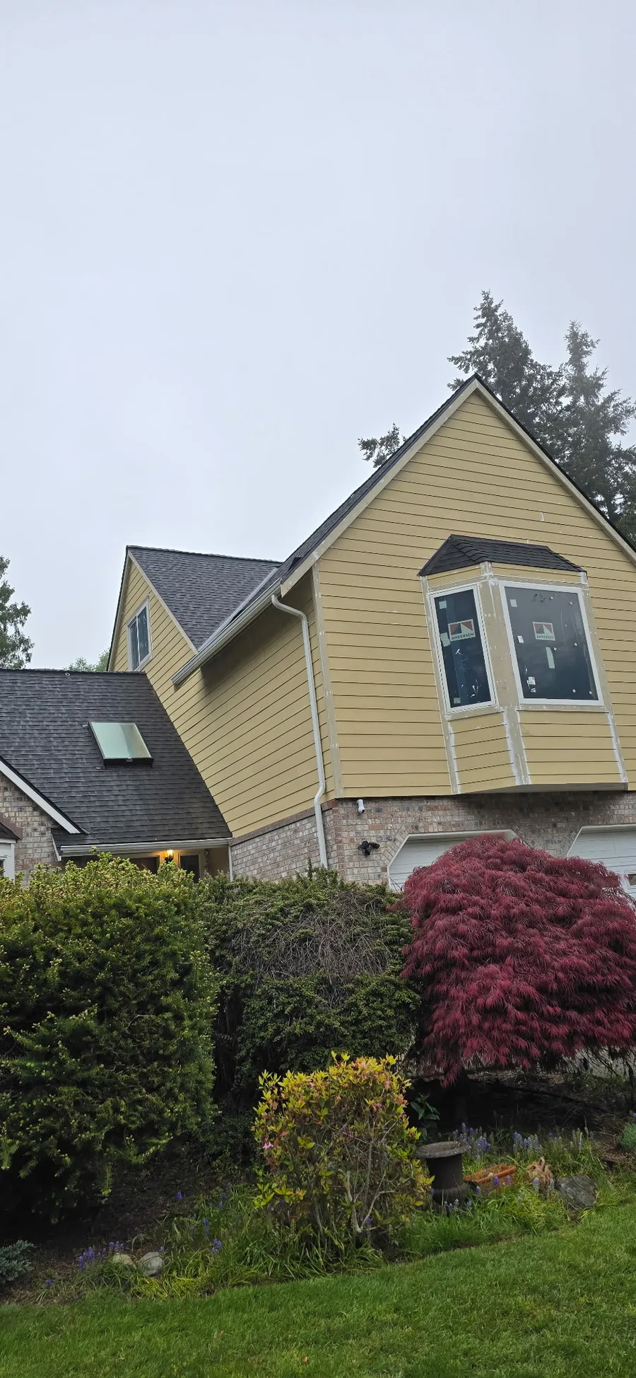 Yellow lap siding on a two-story home with a bay window and brick base, trees and shrubs in front