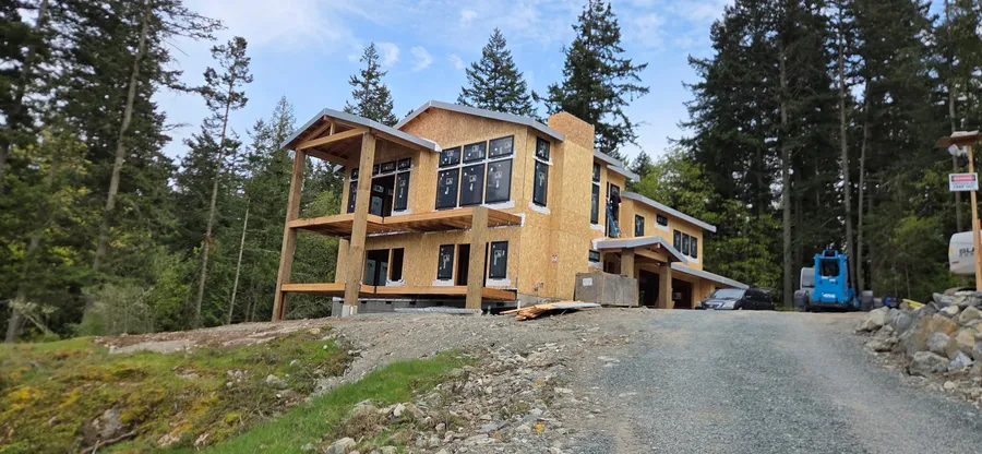 Newly framed home wrapped in OSB sheathing with a timber porch, set among evergreens at the end of a gravel drive