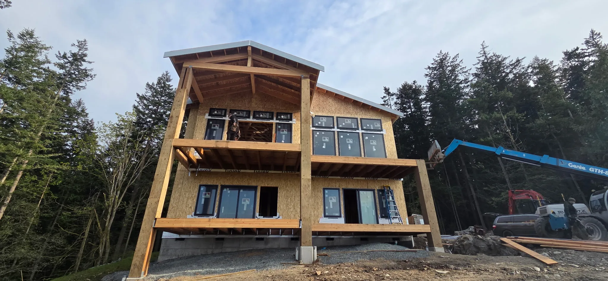 Three-story framed home on a wooded lot with timber posts and OSB sheathing, with a telehandler working beside it