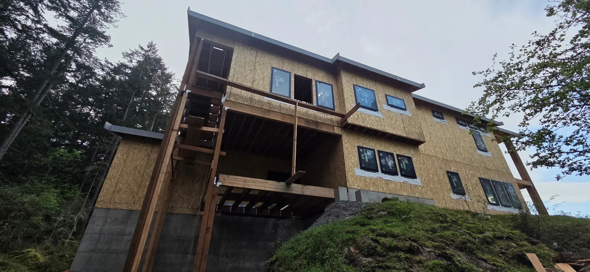Hillside home framed on a concrete foundation with OSB sheathing, windows set, and timber deck framing