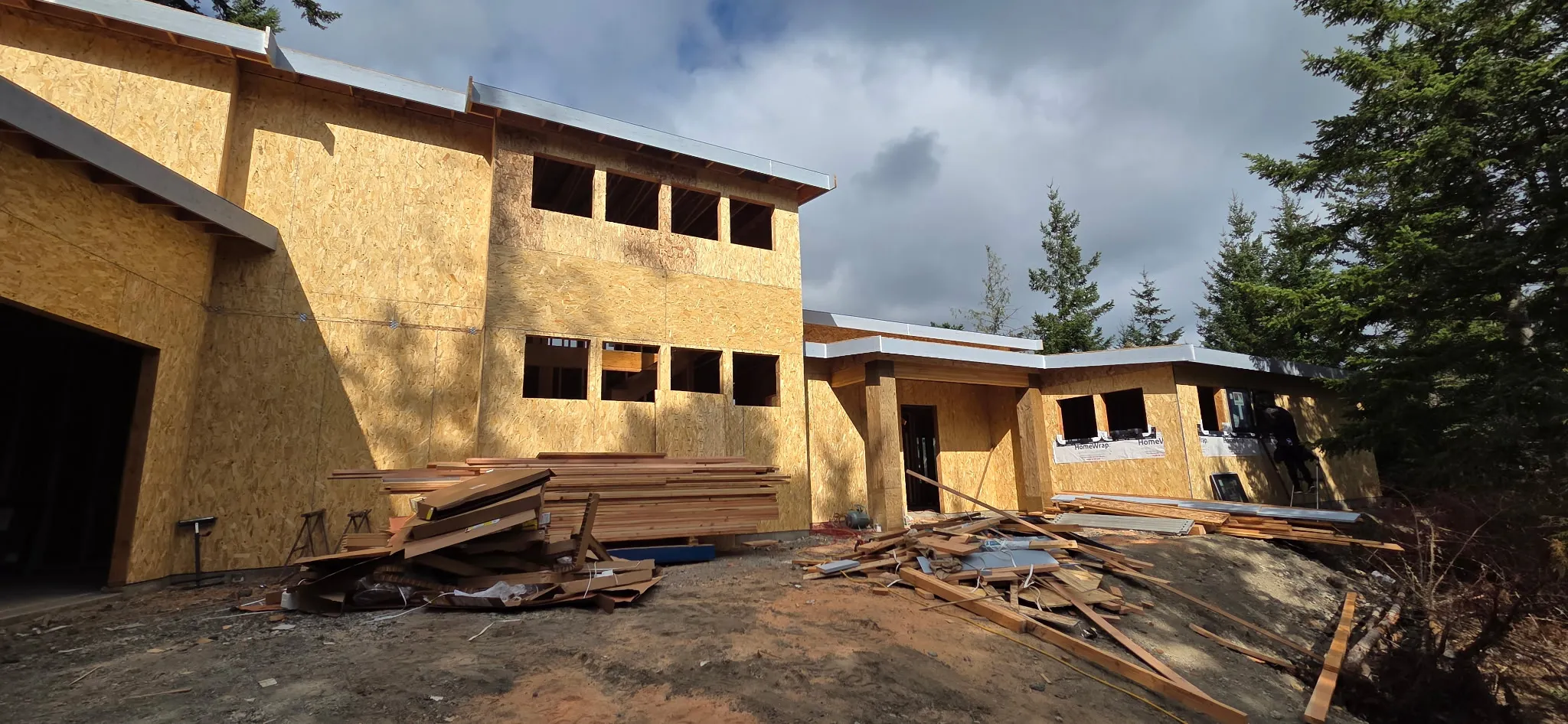 Large single-level framed home with OSB-sheathed walls, window openings, and stacked lumber out front
