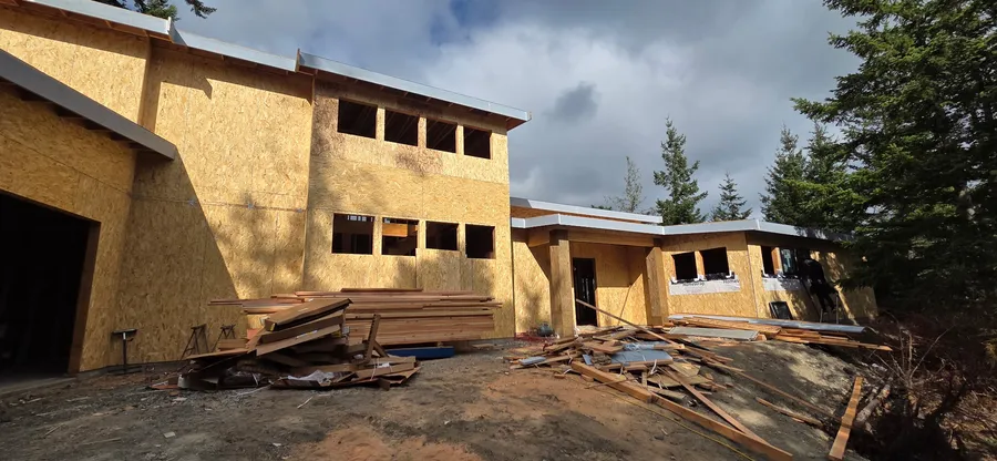 Large single-level framed home with OSB-sheathed walls, window openings, and stacked lumber out front